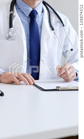 Unreckognisable doctor man writing a prescription and filling out medical records on a clipboard in an office setting. Medicine and health care concept 128874312