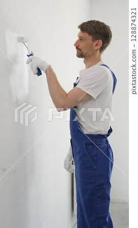 Man professional painter wearing protective white gloves and blue work overalls, applying paint with roller on pristine interior wall during home renovation project, vertical view Man professional painter wearing protective white gloves and blue work overalls, applying paint with roller on pristine interior wall during home renovation project, vertical view 128874321