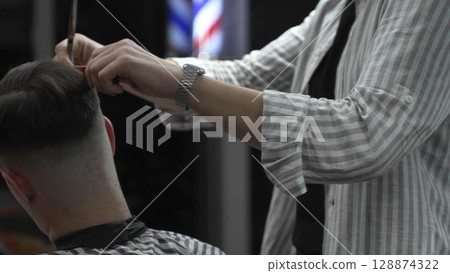 Skilled barber trimming customer's hair using professional scissors and comb, working in contemporary barbershop with vintage pole visible near workspace Skilled barber trimming customer's hair using professional scissors and comb, working in contemporary barbershop with vintage pole visible near workspace 128874322