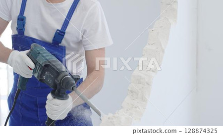 Male construction worker demolishing white wall with rotary hammer drill, wearing blue overalls and protective white gloves, closeup view 128874325
