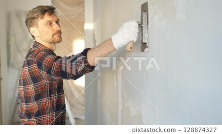 Male construction worker wearing protective gloves smoothing plaster onto wall surface during home renovation using professional trowel tool 128874327