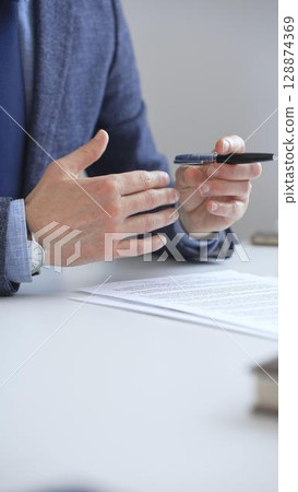Professional businessman gesturing while discussing strategy, seated at white desk with tablet, eyeglasses, and wristwatch reflecting on sleek workspace surface. Business people concept 128874369