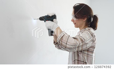 Middle aged female professional worker wearing protective gear and gloves, is drilling hole in white wall during home improvement project, focused on precise home renovation work Middle aged female professional worker wearing protective gear and gloves, is drilling hole in white wall during home improvement project, focused on precise home renovation work 128874392