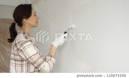 Middle aged woman wearing protective gloves painting white wall with paint roller, updating interior and giving room clean, modern look during home renovation project 128874398