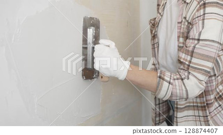 Unknown female construction worker wearing protective gloves applying plaster, smoothing wall surface during home renovation work Unknown female construction worker wearing protective gloves applying plaster, smoothing wall surface during home renovation work 128874407