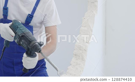 Male construction worker demolishing white wall with rotary hammer drill, wearing blue overalls and protective white gloves, closeup view 128874410