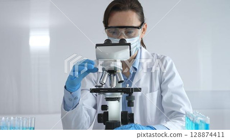 A female scientist in a lab coat and blue gloves examines a microscope slide in a sterile laboratory, conducting research using blue samples from a test tube 128874411