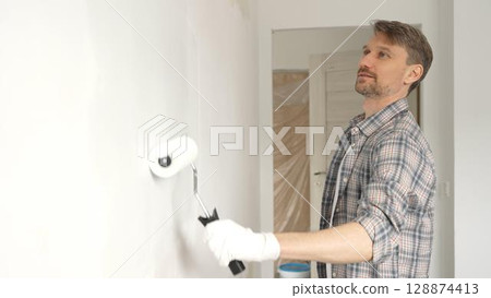 Middle aged man wearing checkered shirt, is rolling white paint on apartment wall with paint roller, transforming interior space with steady strokes during comprehensive home renovation Middle aged man wearing checkered shirt, is rolling white paint on apartment wall with paint roller, transforming interior space with steady strokes during comprehensive home renovation 128874413