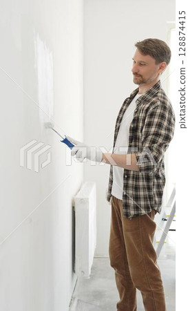 Professional house painter wearing gloves painting a white wall with a paint roller, working on a home renovation project, creating a fresh and clean look 128874415
