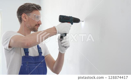 Middle aged man wearing blue overall, is drilling hole in white wall, wearing protective gear during home improvement project with cordless power tool 128874417
