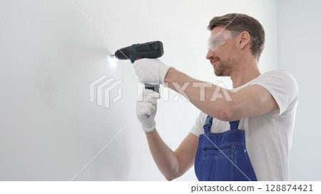 Middle aged man wearing blue overall and protective glasses, is drilling hole in white wall, wearing protective gear during home improvement project with cordless power tool 128874421