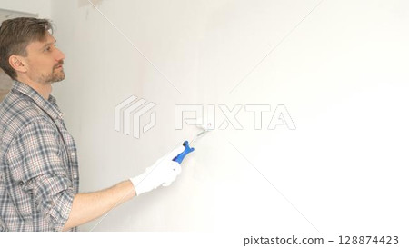 Man professional painter wearing checkered shirt, is rolling white paint on apartment wall, transforming interior space with steady strokes during comprehensive home renovation 128874423