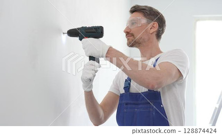 Middle aged man wearing blue overall and protective glasses, is drilling hole in white wall, wearing protective gear during home improvement project with cordless power tool 128874424