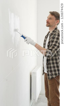 Middle aged man wearing gloves is painting a white wall with a paint roller, working on a home renovation project, creating a fresh and clean look 128874429