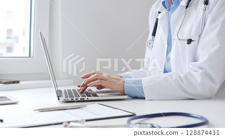 Female doctor is typing on a laptop with a stethoscope, medical record, and pen on the desk in a bright medical office, showcasing healthcare technology. Medicine concept Female doctor is typing on a laptop with a stethoscope, medical record, and pen on the desk in a bright medical office, showcasing healthcare technology. Medicine concept 128874431