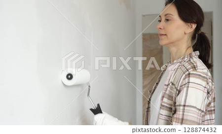 Middle aged woman wearing gloves is skillfully applying paint to a white wall with a paint roller in an apartment undergoing renovation, transforming the space with her creative touch Middle aged woman wearing gloves is skillfully applying paint to a white wall with a paint roller in an apartment undergoing renovation, transforming the space with her creative touch 128874432