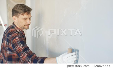 Construction worker man wearing gloves and holding a putty knife is applying plaster on a white wall, performing home renovation and improvement Construction worker man wearing gloves and holding a putty knife is applying plaster on a white wall, performing home renovation and improvement 128874433