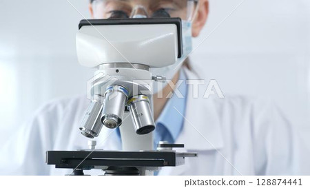 Scientist wearing lab coat and protective glasses using a modern microscope in a laboratory setting, conducting research and experiments in a scientific environment. Health care and science concept 128874441