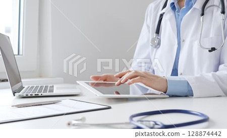 Female doctor wearing white medical coat and stethoscope using digital tablet with laptop and medical records on desk in medical office, showcasing modern healthcare technology. Medicine and helthcare Female doctor wearing white medical coat and stethoscope using digital tablet with laptop and medical records on desk in medical office, showcasing modern healthcare technology. Medicine and helthcare 128874442