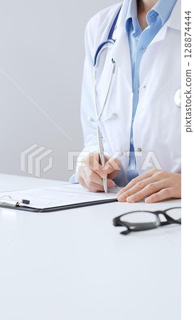Unknown female doctor completing healthcare paperwork, clipboard positioned near laptop and stethoscope in well lit clinical workspace by window. Medicine and health care 128874444