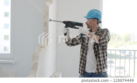 Man construction worker demolishing a wall with a hammer drill in protective gear, creating construction debris on the surrounding floor surface. Renovation concept Man construction worker demolishing a wall with a hammer drill in protective gear, creating construction debris on the surrounding floor surface. Renovation concept 128874449