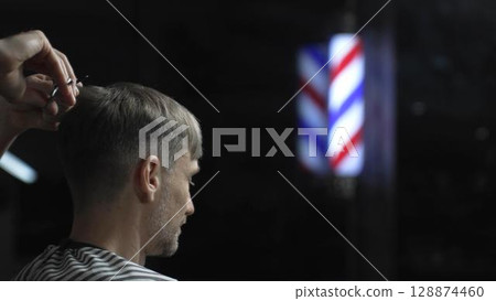 Professional barber grooming male client's hair with scissors, vintage barber pole framing traditional barbershop setting 128874460