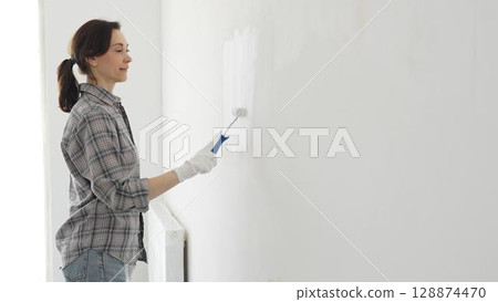 Middle aged woman wearing plaid shirt and white gloves is painting white wall with paint roller, experiencing home renovation and transforming living space. Home repair concept Middle aged woman wearing plaid shirt and white gloves is painting white wall with paint roller, experiencing home renovation and transforming living space. Home repair concept 128874470