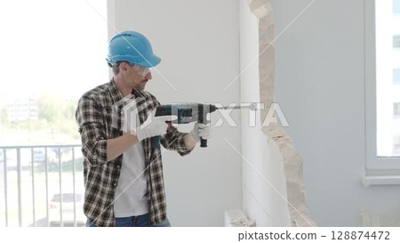 Man construction worker demolishing a wall with a hammer drill in protective gear, creating construction debris on the surrounding floor surface. Renovation concept 128874472
