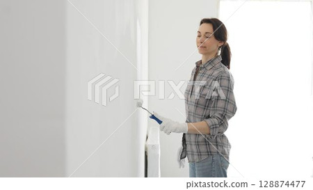 Middle aged happy woman wearing checked shirt and white gloves is painting white wall with paint roller, experiencing home renovation and transforming living space 128874477