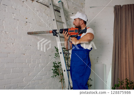 Individual in blue overalls operating a power drill on an indoor white brick wall 128875986