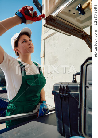Woman in green overalls fixing outdoor air conditioning unit 128875993