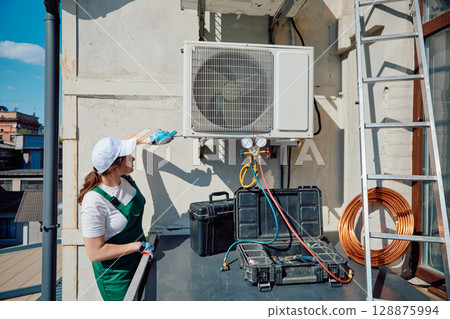 Technician inspecting air conditioning unit on building rooftop under clear blue sky 128875994
