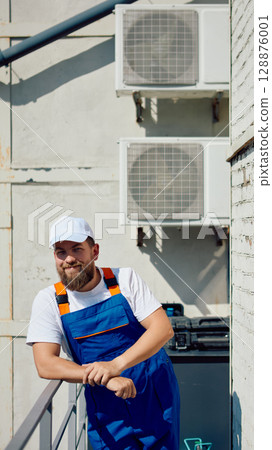 Smiling technician in blue overalls with air conditioner units in background 128876001