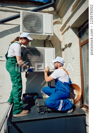 Technicians wearing overalls set up external air conditioning units on building 128876002