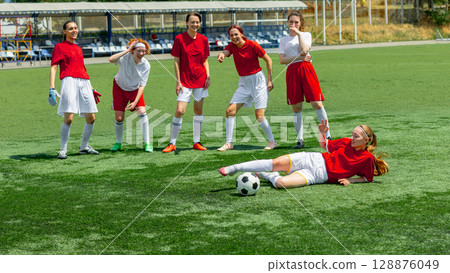 Smiling female footballers standing and joking around fallen teammate 128876049