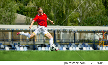 Woman football player in red jersey kicking soccer ball on green field 128876053