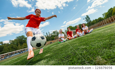 Young women footballer kicks ball while teammates watch on sunny field day Young women footballer kicks ball while teammates watch on sunny field day 128876056
