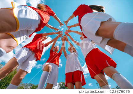 Girls football team in red and white forming team huddle with hands raised under blue sky 128876076