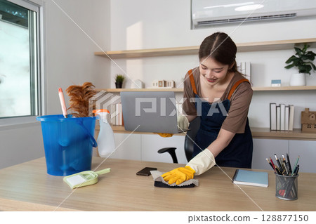 Home Cleaning. Young woman happily organizing her workspace while cleaning. Home Cleaning. Young woman happily organizing her workspace while cleaning. 128877150