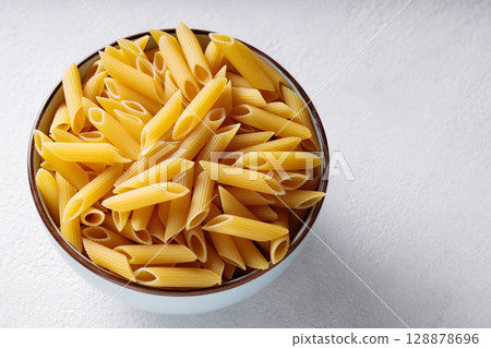 Raw Penne Pasta in Ceramic Bowl on White Background Copy Space 128878696
