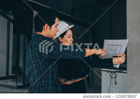 Construction manager and engineer dressed in orange work vests and hard helmets explore construction documentation on the building site near the steel frames Construction manager and engineer dressed in orange work vests and hard helmets explore construction documentation on the building site near the steel frames 128878807
