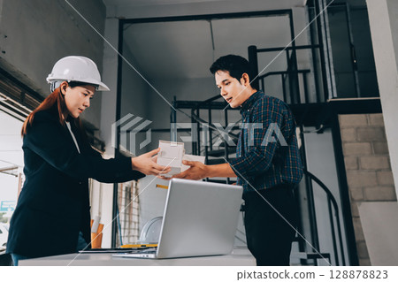 Construction manager and engineer dressed in orange work vests and hard helmets explore construction documentation on the building site near the steel frames 128878823