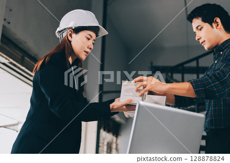 Construction manager and engineer dressed in orange work vests and hard helmets explore construction documentation on the building site near the steel frames 128878824