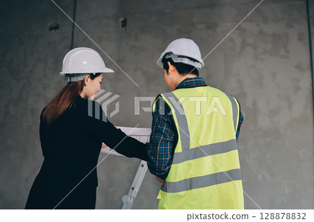 Construction manager and engineer dressed in orange work vests and hard helmets explore construction documentation on the building site near the steel frames 128878832