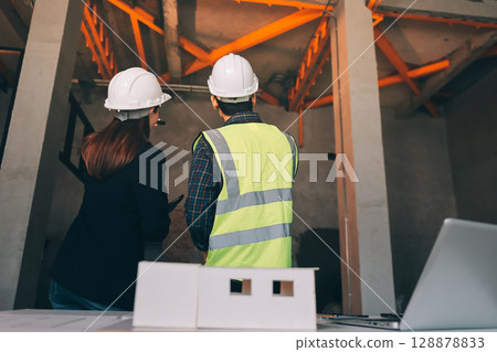 Construction manager and engineer dressed in orange work vests and hard helmets explore construction documentation on the building site near the steel frames 128878833
