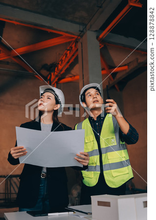 Construction manager and engineer dressed in orange work vests and hard helmets explore construction documentation on the building site near the steel frames 128878842