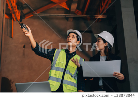 Construction manager and engineer dressed in orange work vests and hard helmets explore construction documentation on the building site near the steel frames Construction manager and engineer dressed in orange work vests and hard helmets explore construction documentation on the building site near the steel frames 128878844