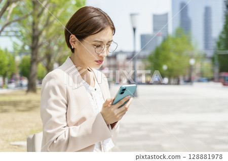 一位面帶愁容的年輕女商人坐在長椅上,操作著智慧型手機 一位面帶愁容的年輕女商人坐在長椅上,操作著智慧型手機 128881978