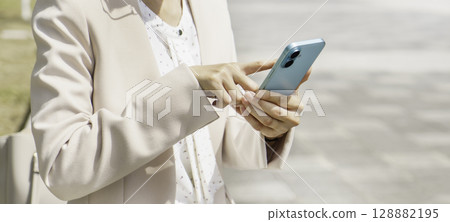 Close-up of a young businesswoman sitting on a bench and operating her smartphone, banner size Close-up of a young businesswoman sitting on a bench and operating her smartphone, banner size 128882195