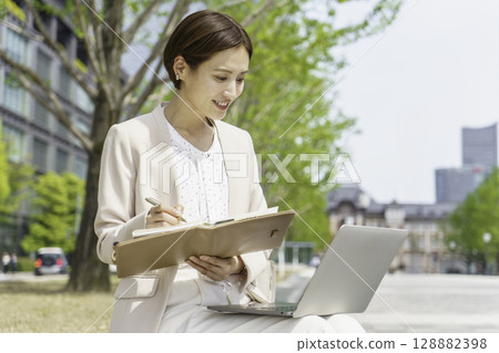 A young businesswoman sitting on a bench and writing in her notebook while looking at her laptop A young businesswoman sitting on a bench and writing in her notebook while looking at her laptop 128882398